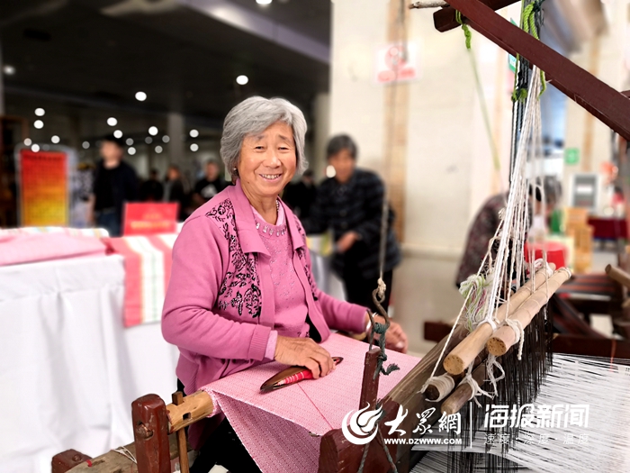 在首届菏泽扶贫车间产品展销会上,杨桂芝老人坐在一台织布机上织鲁锦